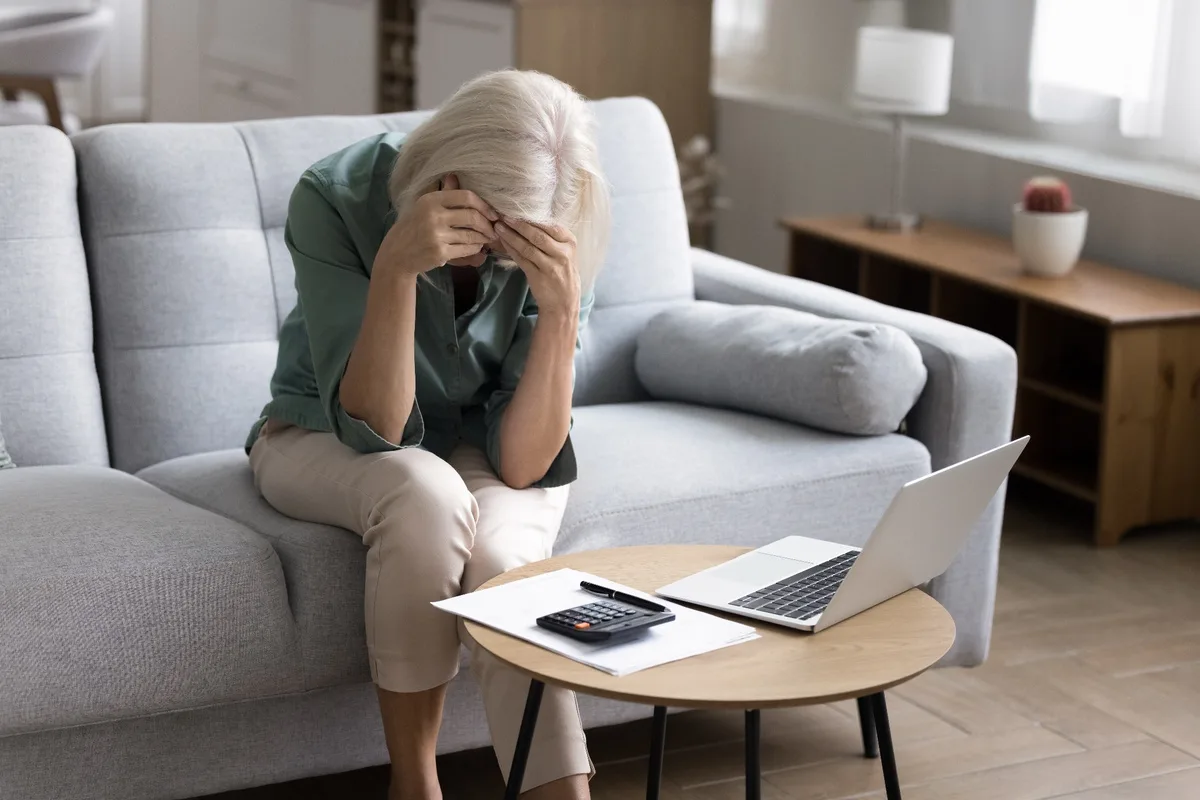 Mature woman sitting with her head in her hands next to a laptop and a calculator